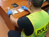 Na zdjęciu policjant w kamizelce odblaskowej w kolorze żółtym wykonuje czynności z leżącymi na biurku narkotykami znajdującymi się w foliowych woreczkach, obok nich widoczna elektroniczna waga.