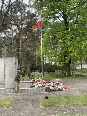 widoczny pomnik na cmentarzu wojskowym przy pomniku widać wiązanki kwiatów oraz maszt z flaga państwowa  biało - czerwoną
