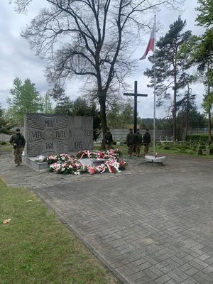 widoczny pomnik na cmentarzu wojskowym pod pomnikiem wiązanki kwiatów po obu stronach pomnika stoją żołnierze