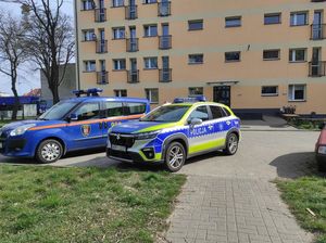 na zdjęciu widoczny jest oznakowany radiowóz stojący obok oznakowanego samochodu straży miejskiej na parkingu przed blokiem